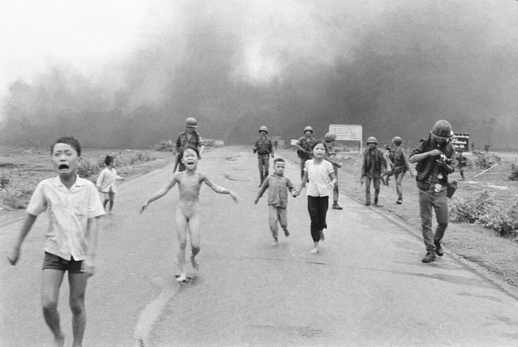Fotografía en blanco y negro de una niña desnuda corriendo por una carretera con los brazos abiertos, con humo de fondo, durante la guerra de Vietnam
